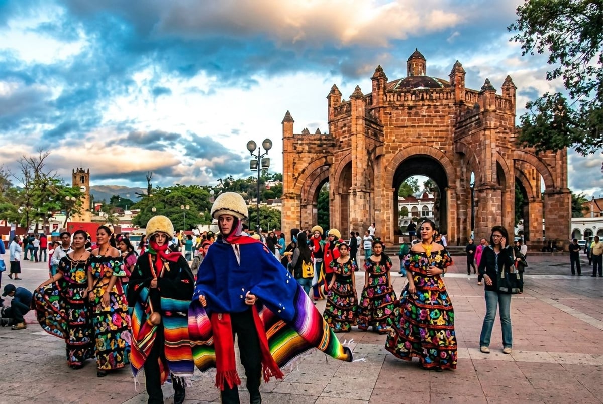 Colorful performers in traditional Mexican clothing and decorated costumes in front of an ornate stone archway in a town plaza
