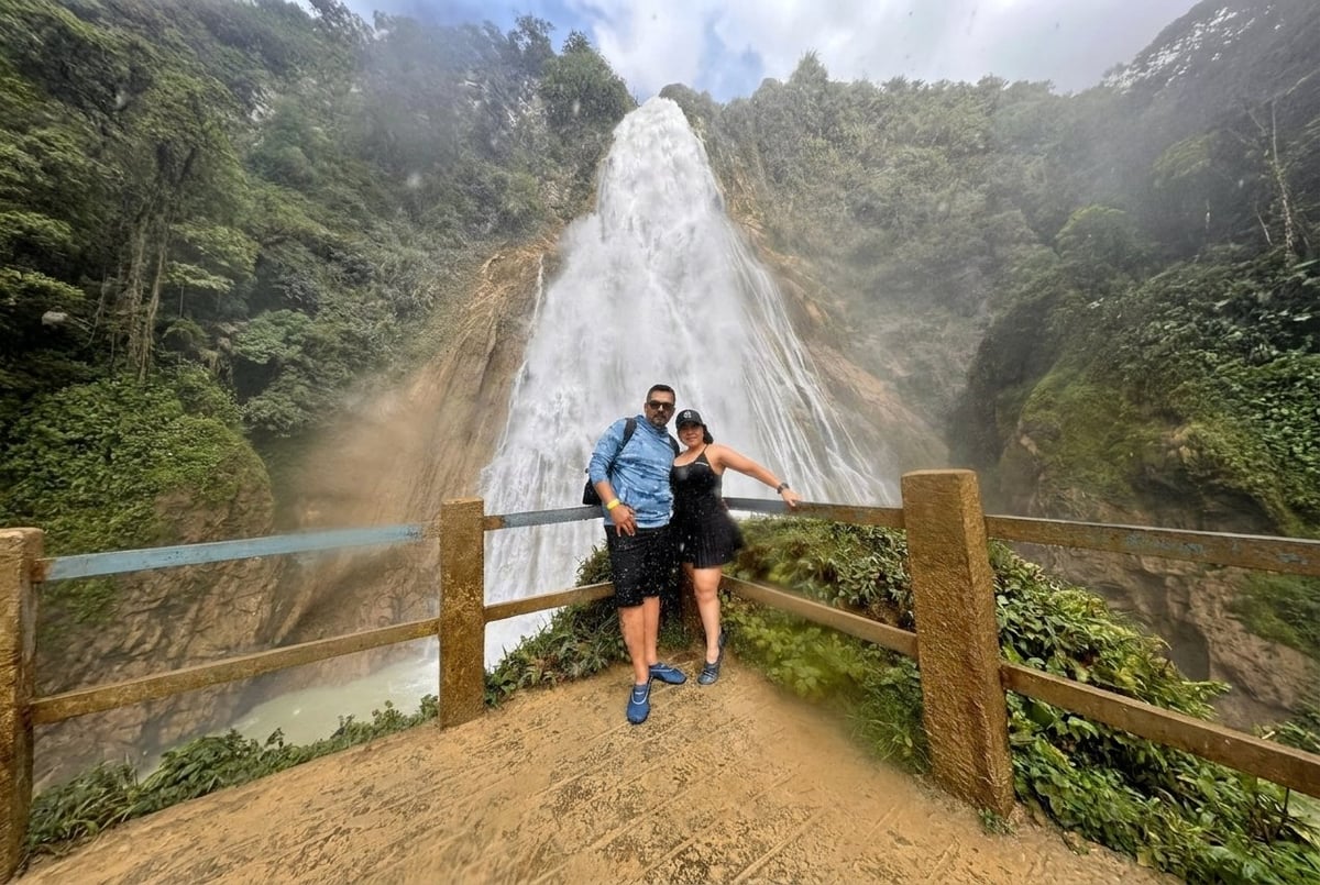 Two hikers stand at a wooden railing overlooking a tall waterfall cascading through lush green mountains
