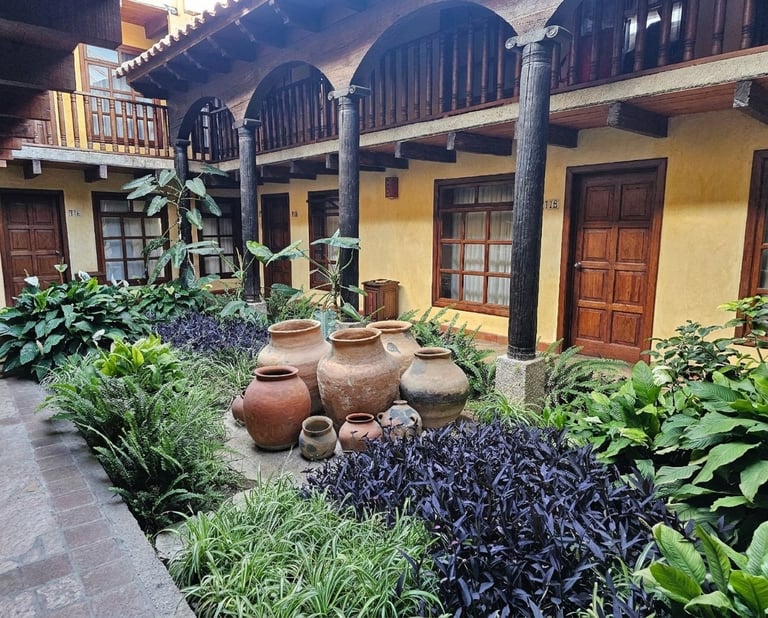 Colonial courtyard with yellow stucco walls, wooden arches, terracotta pots, and lush vegetation surrounding a central garden area