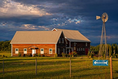 Red barn with tan roof and attached wooden windmill beside a fenced pasture under dramatic blue sky