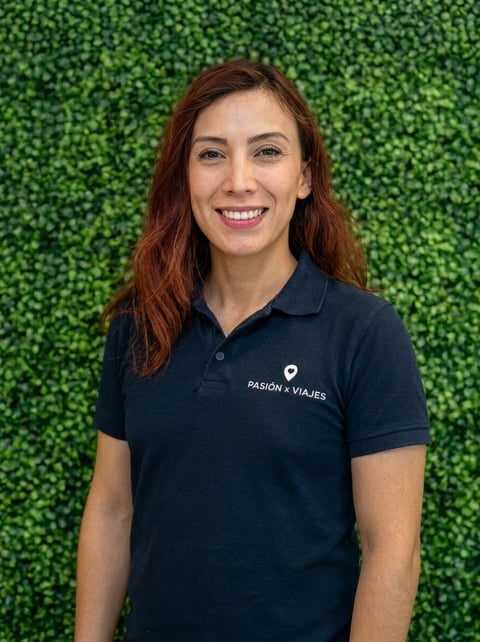 Woman with reddish-brown hair smiling at camera wearing dark polo shirt with Pasión x Viajes logo against green leafy background