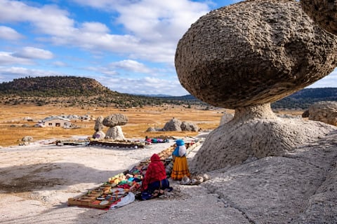 Woman in red jacket sits beneath massive balanced boulder overlooking valley settlement with mountains in background